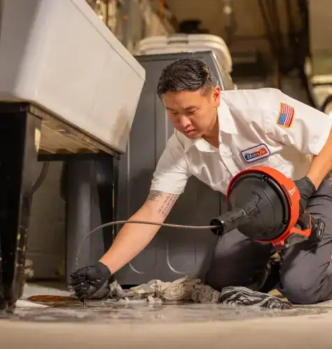 A Bonfe plumber kneels on the floor in an indoor utility area, professionally clearing a clog with a drain snake. He wears protective gloves and a Bonfe uniform shirt featuring the company patch, providing expert service to keep your plumbing running smoothly.