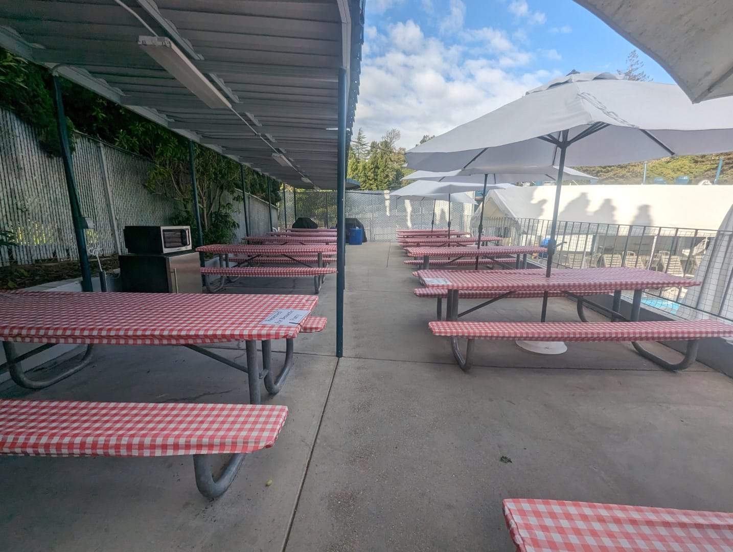 Empty outdoor picnic tables with red checkered tablecloths at Bay-O-Vista Swim & Tennis Club, featuring shaded seating areas and umbrellas, ideal for relaxing and socializing.