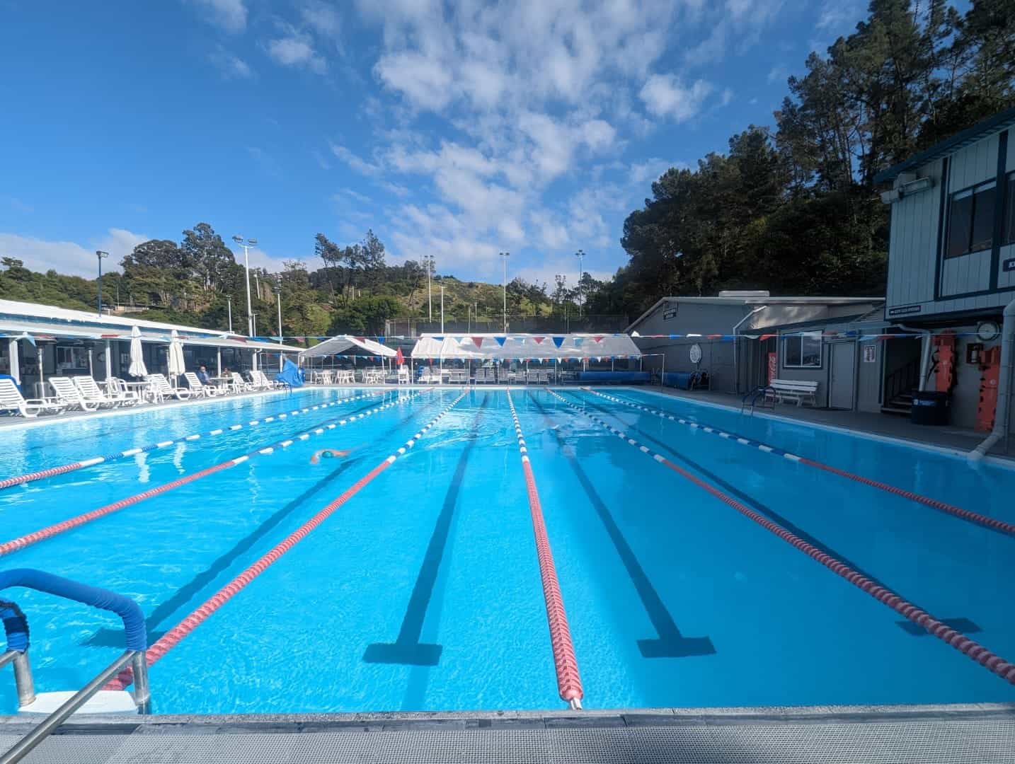 Bright outdoor swimming pool at Bay-O-Vista Swim & Tennis Club, surrounded by lounge chairs and shaded areas, with a scenic hillside and clear blue sky in the background.