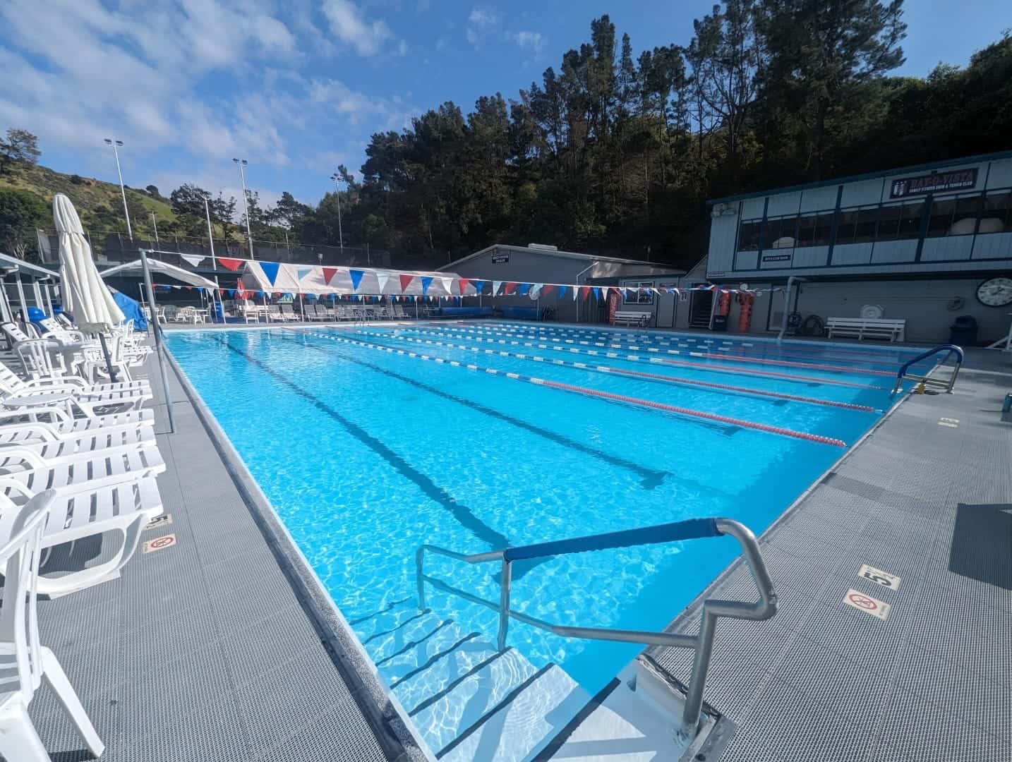 Bright outdoor swimming pool at Bay-O-Vista Swim & Tennis Club, featuring multiple lanes, swimming flags, and surrounding lounge chairs on a sunny day.