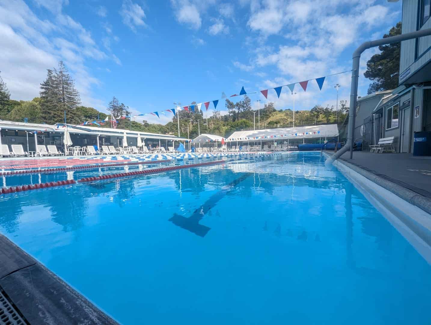 Bright outdoor swimming pool at Bay-O-Vista Swim & Tennis Club with blue water, pool lanes, and colorful pennant banners under a partly cloudy sky.