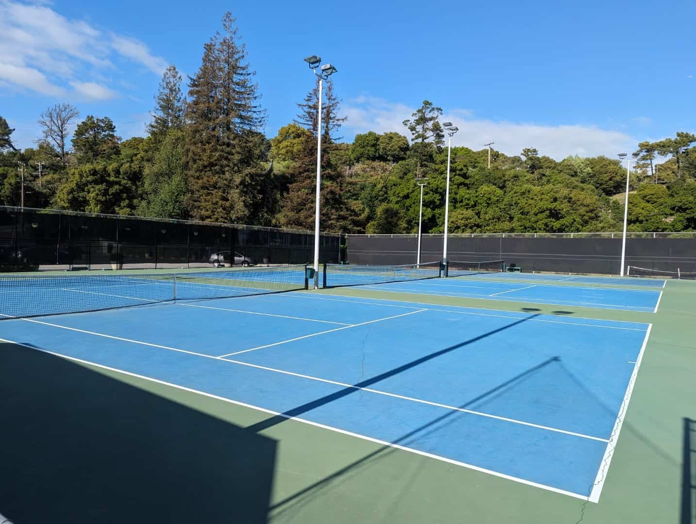 Bright blue outdoor tennis courts at Bay-O-Vista Swim & Tennis Club surrounded by lush green trees under clear blue skies.