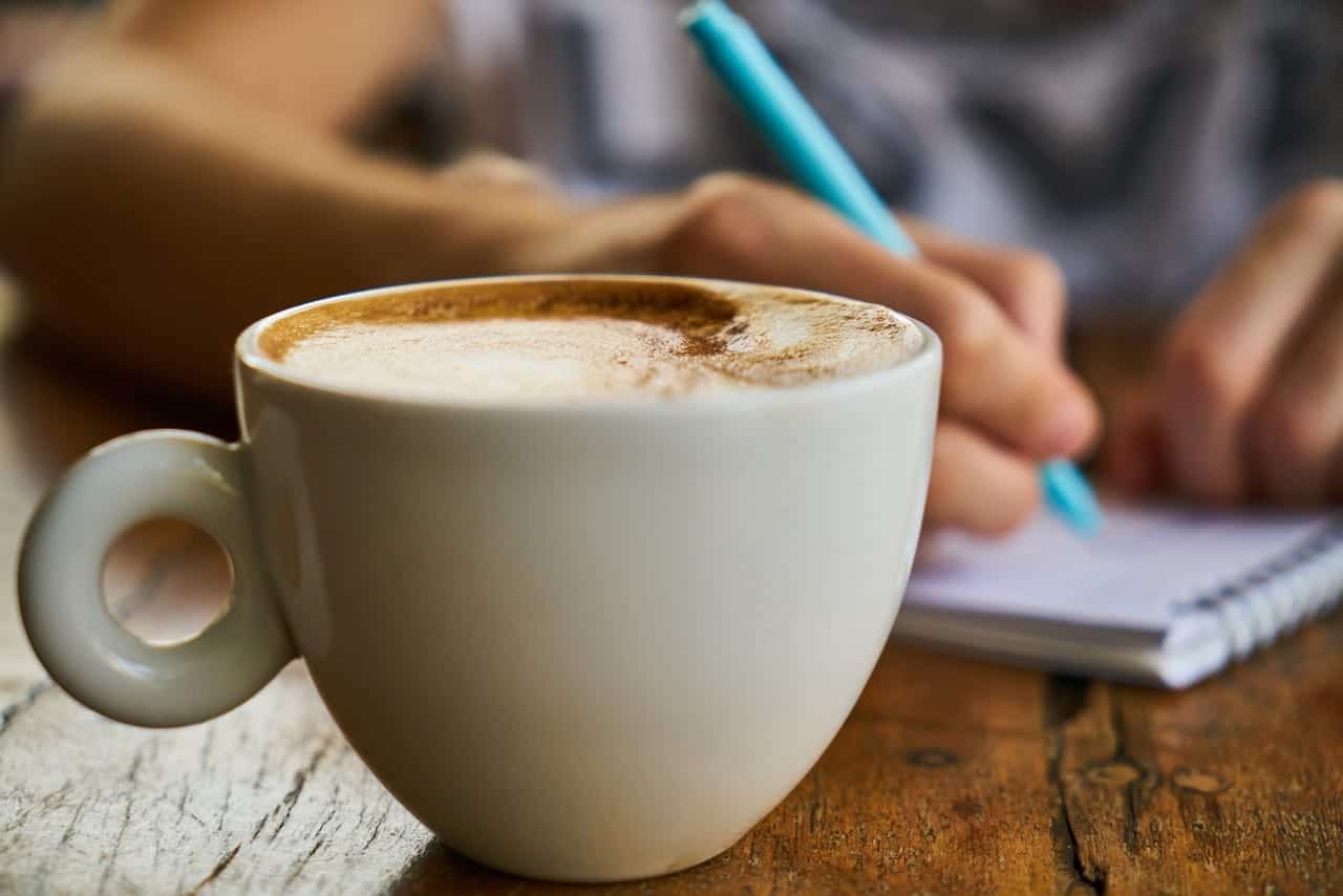 A person taking notes with a blue pen beside a cup of frothy cappuccino on a wooden table at Bay-O-Vista Swim & Tennis Club.