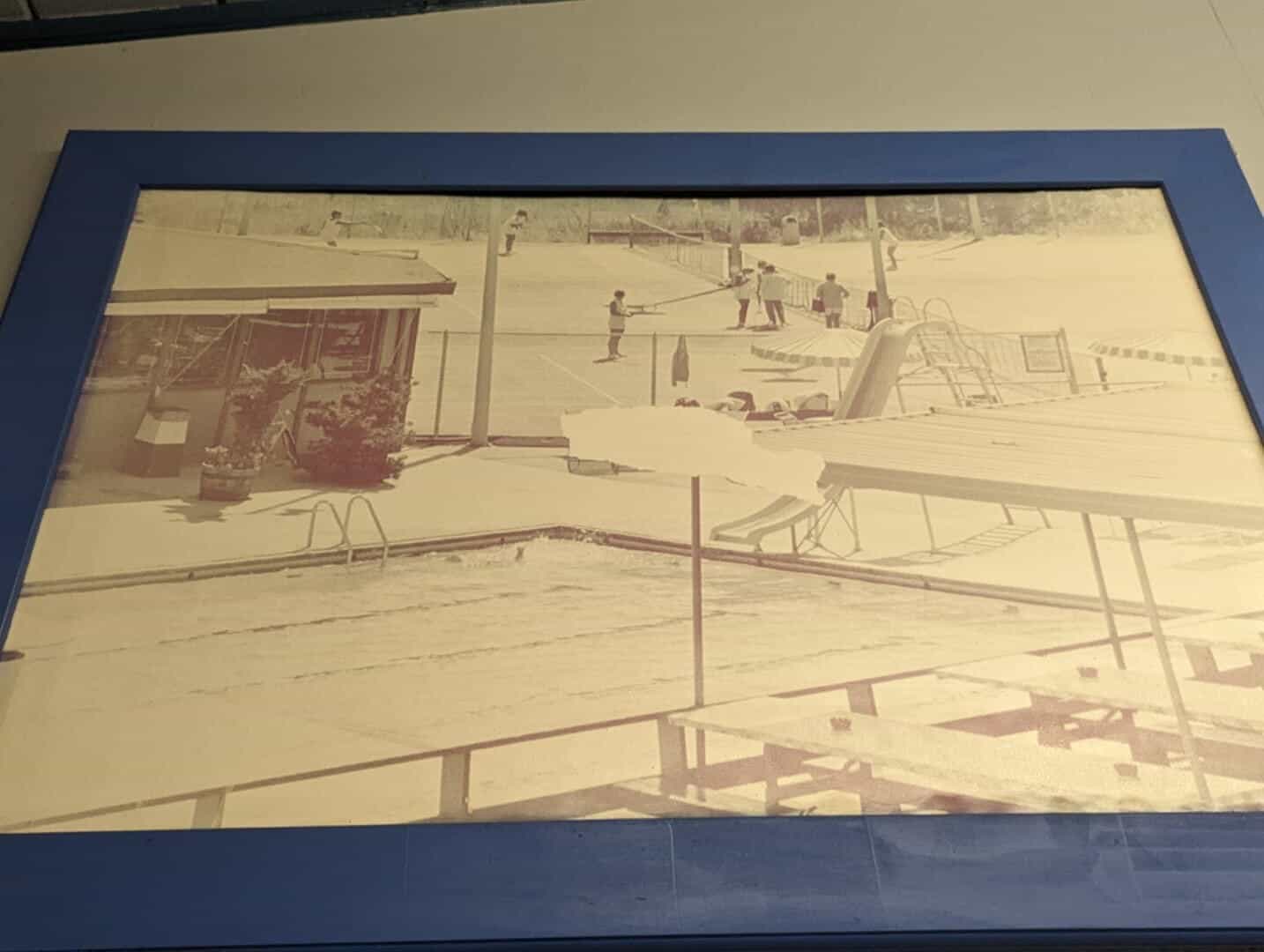 A vintage black and white photograph of the Bay-O-Vista Swim & Tennis Club showing an outdoor swimming pool with a slide and children playing on the court, highlighting community recreational facilities.