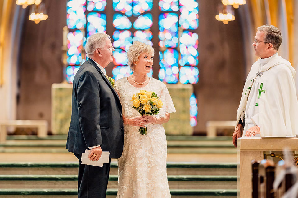 An older couple dressed formally stands at the altar with a bouquet of yellow roses, speaking to a priest inside the brightly lit St Joseph the Workman Cathedral in La Crosse, with stained glass windows gleaming.