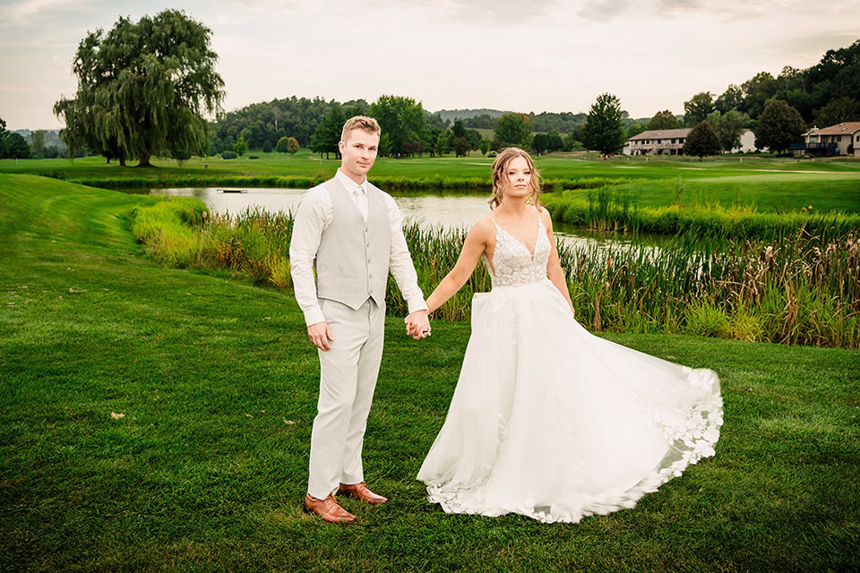 fox-hollow-wedding-bride-and-groom-walking.jpg A bride and groom stand holding hands on a grassy lawn near a pond at a Fox Hollow wedding in La Crosse, dressed in wedding attire with trees and buildings in the background.