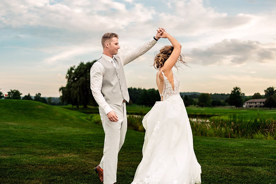 fox-hollow-wedding-couple-twirl-at-sunset.jpg A bride and groom dance on a grassy field at their Fox Hollow wedding in La Crosse, with the groom spinning the bride under his arm; a cloudy sky and trees are in the background.