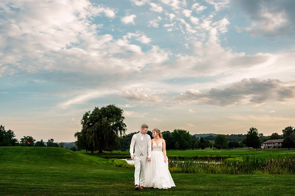 fox-hollow-wedding-wide-landscape-photo.jpg A bride and groom walk hand in hand on a grassy field at their Fox Hollow wedding in La Crosse, with trees and a pond in the background under a partly cloudy sky.