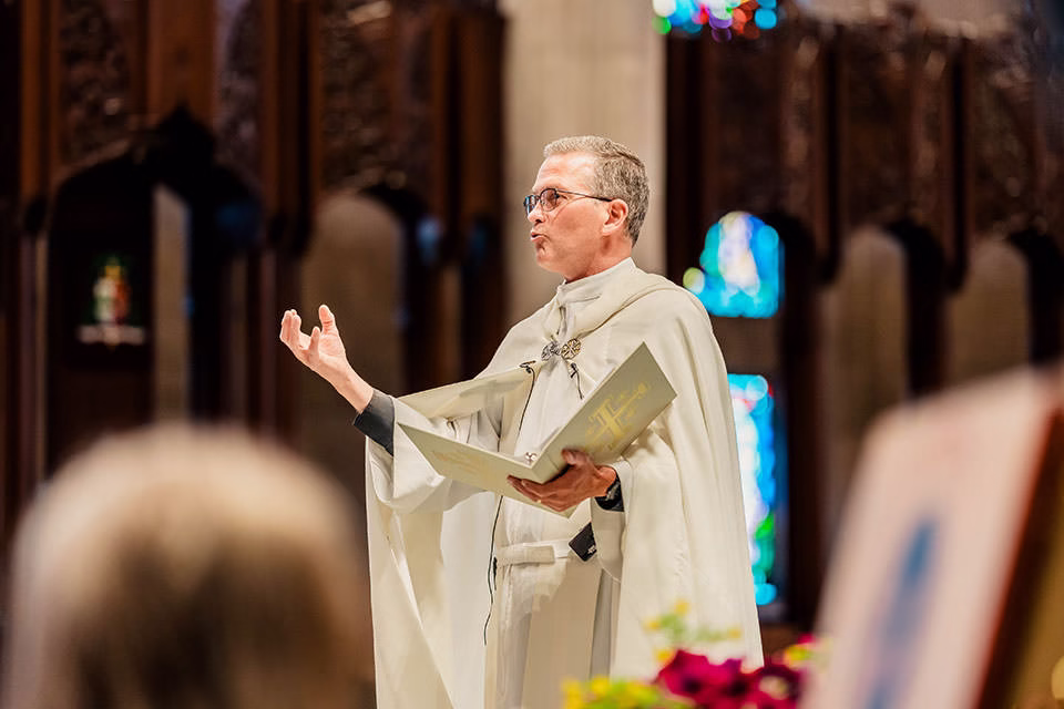 A priest in white robes holds a book and gestures while speaking inside St Joseph the Workman Cathedral in La Crosse, with stained glass windows and wooden columns.