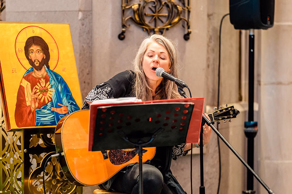 A woman plays an acoustic guitar and sings into a microphone next to a religious icon of Jesus during a St Joseph the Workman Cathedral wedding in La Crosse.