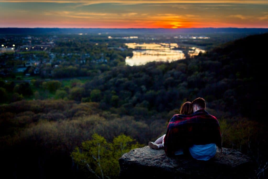 Adventure Couple Photos An adventurous couple captures their engagement photos on a rock, admiring the cityscape during a picturesque sunset.