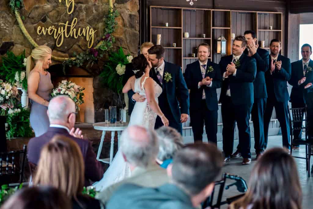 A bride and groom kiss in front of a fireplace during their wedding ceremony.