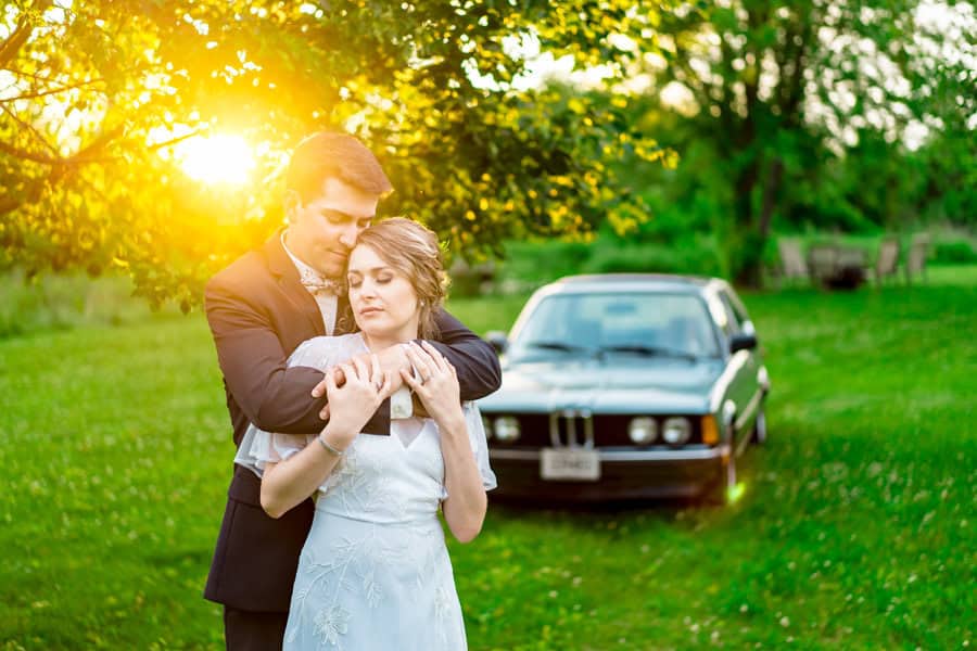 A bride and groom hugging in front of a car.