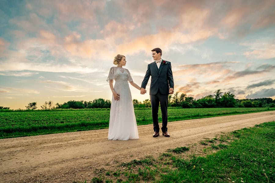 A bride and groom standing on a dirt road at sunset.