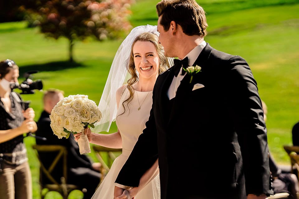 A bride in a white dress and veil holds a bouquet of white flowers, smiling at the groom in a black tuxedo during their outdoor Justin Trails Barn Wedding ceremony.