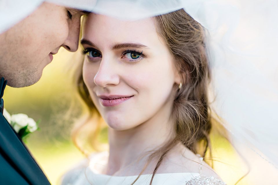 Close-up of a bride smiling at the camera while standing next to the groom, both partially covered by a white veil at their Justin Trails Barn Wedding.