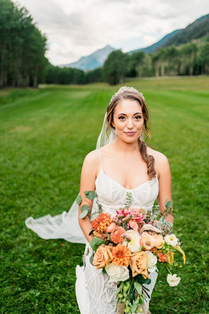 A bride in a white dress and veil stands on green grass, holding a colorful bouquet, with trees and mountains in the background—perfect for a romantic TLazy Ranch Wedding.
