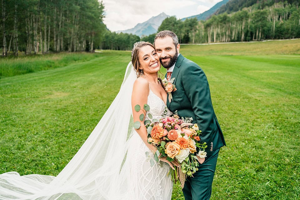 A bride and groom stand close together on a grassy field at their TLazy Ranch Wedding, smiling at the camera. The bride holds a bouquet of flowers, with mountains and trees visible in the background.