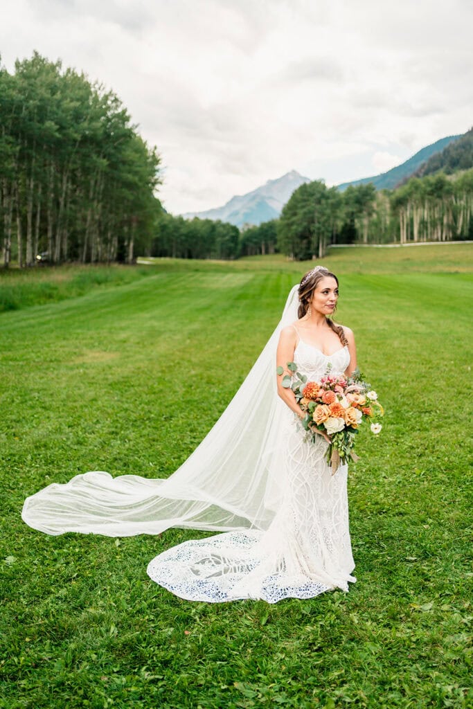 A bride in a white dress and long veil stands on a grassy field at her TLazy Ranch Wedding, holding a large bouquet of flowers with trees and mountains in the background.