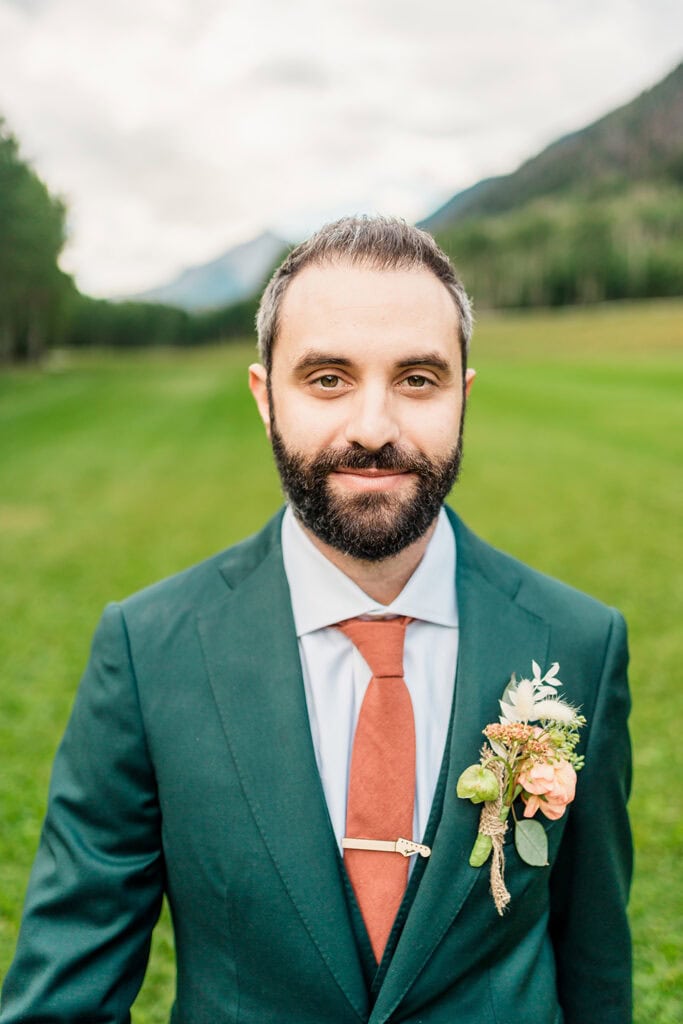 A man with a beard stands outdoors on a grassy field at a TLazy Ranch Wedding, wearing a green suit, rust-colored tie, and floral boutonniere. Trees and mountains are visible in the background.