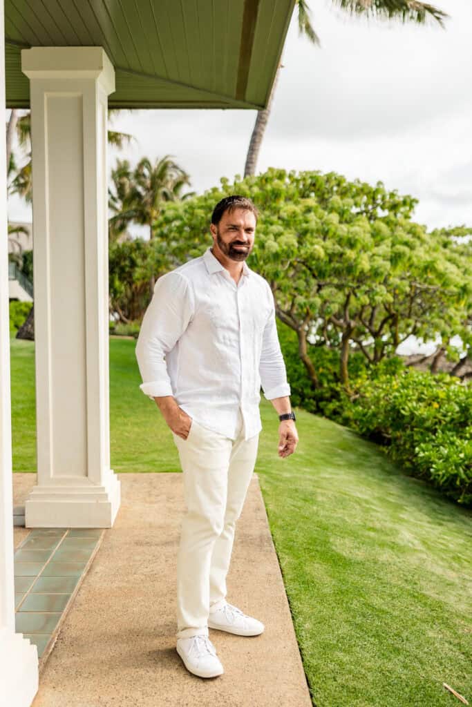 A man in a white shirt and pants stands outdoors on a patio near greenery and a column on a sunny day.