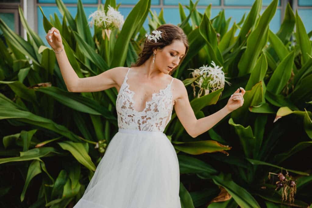 A woman in a white dress with lace details poses with her arms raised in front of large green leaves and white flowers.