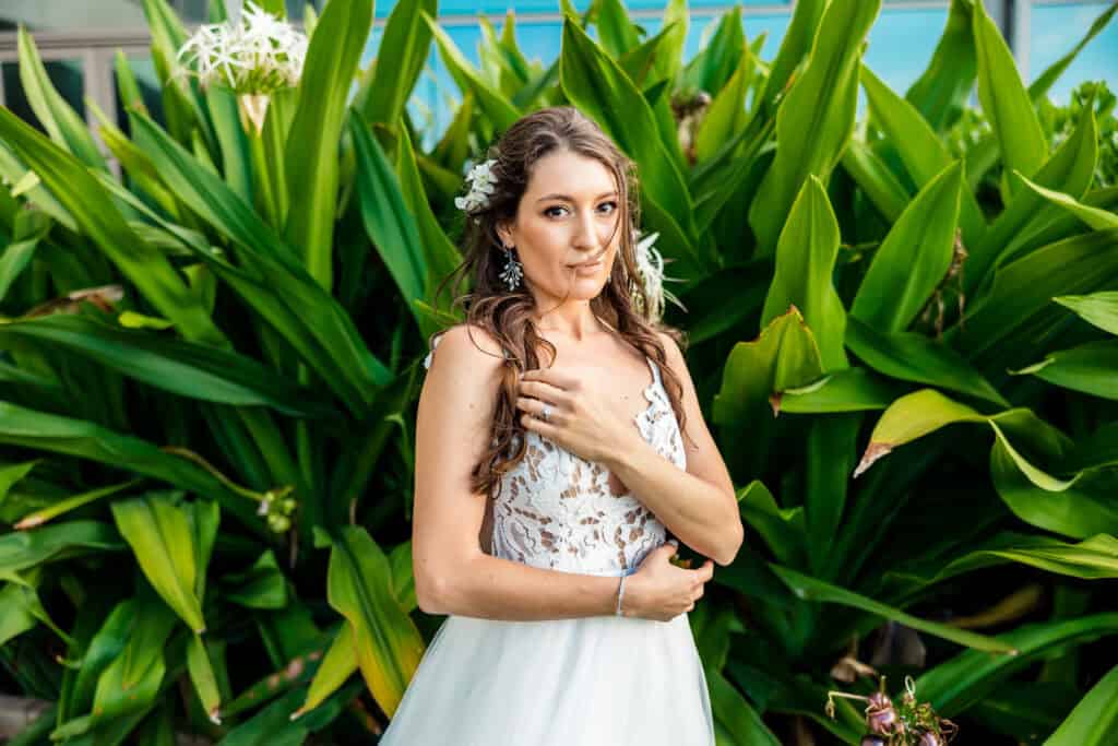 A woman in a white lace dress stands in front of large green plants, holding her right hand to her chest and looking towards the camera. She has flowers in her hair and wears earrings.