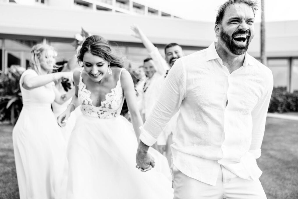A group of people wearing white outfits enjoy a celebratory moment outdoors, with a man and a woman holding hands and laughing in the foreground.
