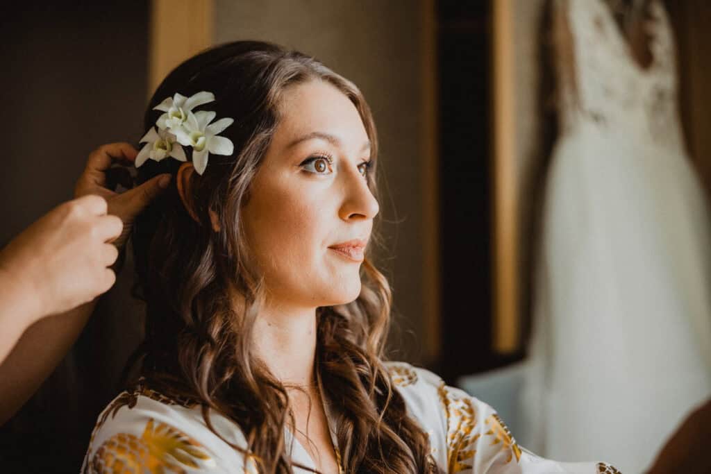 A woman with long hair is being assisted in placing a white flower in her hair. She is wearing a patterned robe.