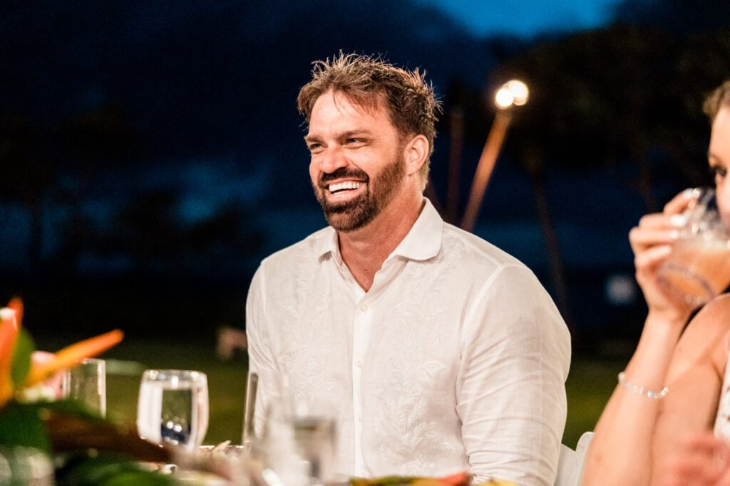 A man in a white shirt is sitting at a table outdoors, smiling and looking to his right. The table has glasses and greenery. The background is dark with trees silhouetted against the sky.