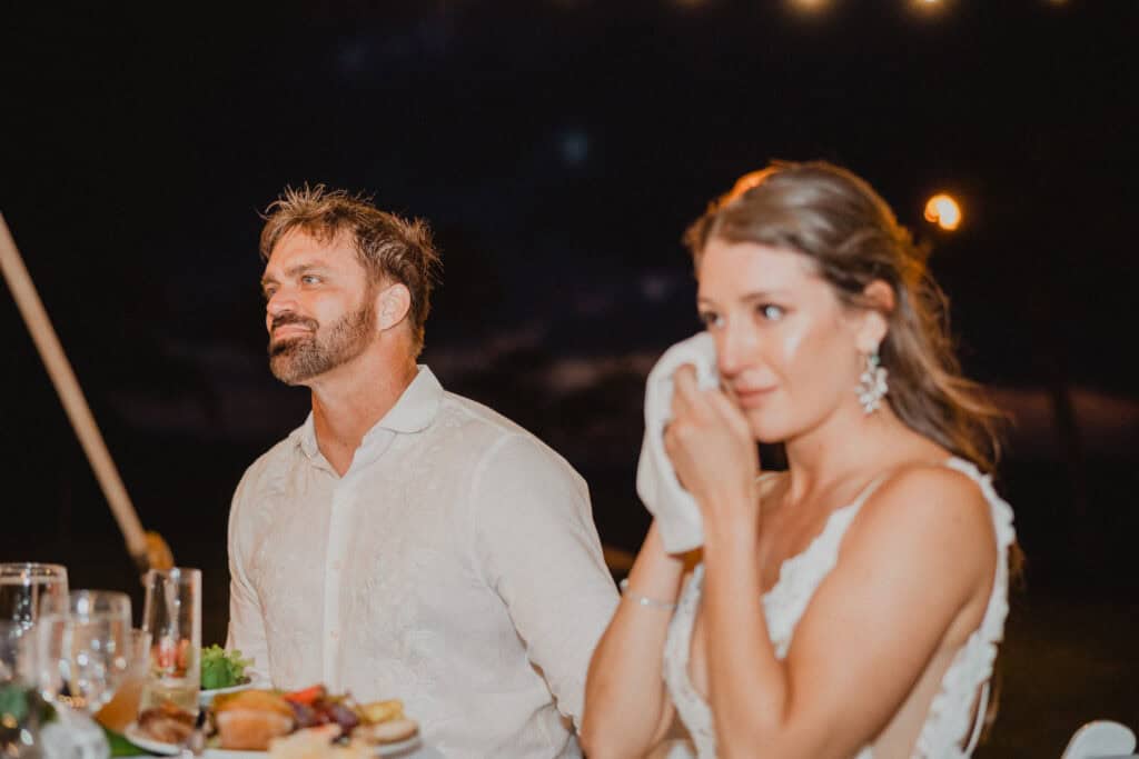 A man and a woman in formal attire are seated at a table outdoors at night. The woman is wiping her tears with a napkin. Plates of food and glasses are visible on the table.