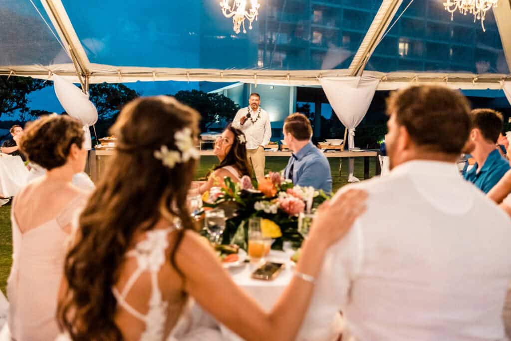 A man stands and makes a speech at a wedding reception under a tent. Guests sit at round tables covered with white tablecloths, with floral centerpieces and food. A chandelier hangs above.
