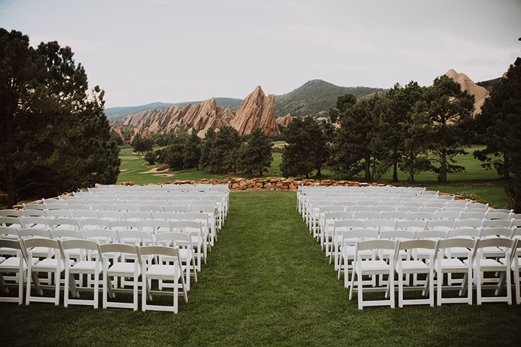Rows of white chairs are arranged outdoors on a lawn at an Arrowhead Golf Club Wedding, facing a scenic view of rocky mountains and trees under a cloudy sky.