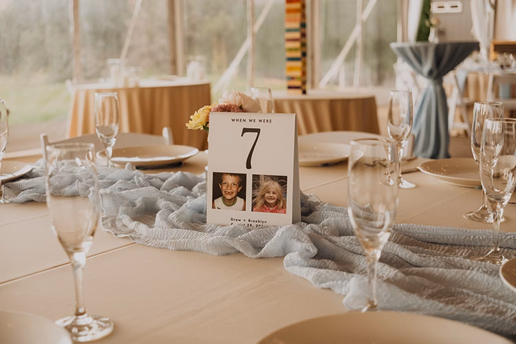 A wedding table setting at an Arrowhead Golf Club Wedding features a numbered card with two childhood photos and a blue table runner, surrounded by plates and glassware.