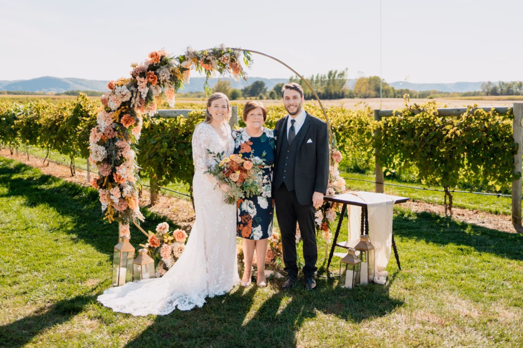 elmaro-vineyard-table-decor A bride, groom, and an older woman stand in front of a floral arch outdoors at a vineyard wedding ceremony on a sunny day.