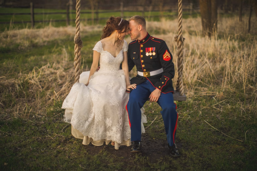 A bride in a white dress and a groom in a military uniform sit closely together on a wooden swing outdoors in a grassy field.