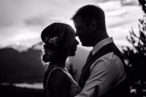A couple stands close together outdoors at sunset, facing each other in silhouette, with distant mountains and trees in the background.