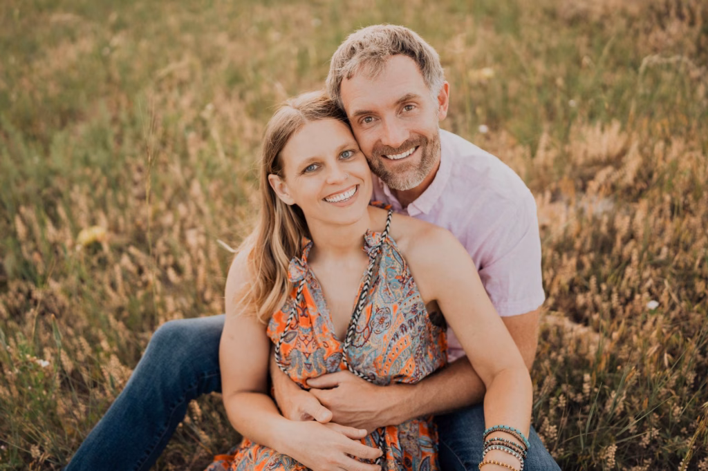A couple sits closely together on grass, both smiling at the camera. The woman wears a patterned dress, and the man wears a light shirt and jeans.