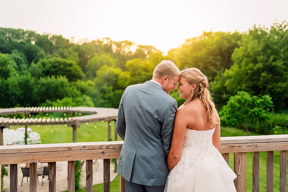 A bride and groom stand close together on a wooden deck, facing away, with a green outdoor setting and sunset in the background at their Barn at Copper Creek Wedding.