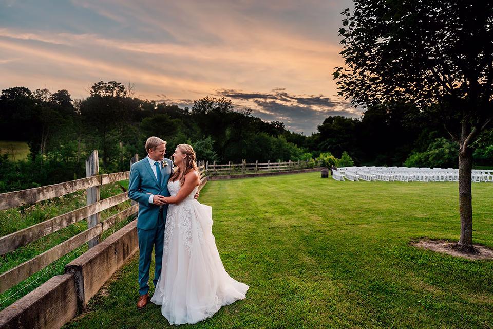 A bride and groom stand together on a grassy lawn near a wooden fence at sunset, with white chairs arranged in the background—a picture-perfect moment at their Barn at Copper Creek Wedding.