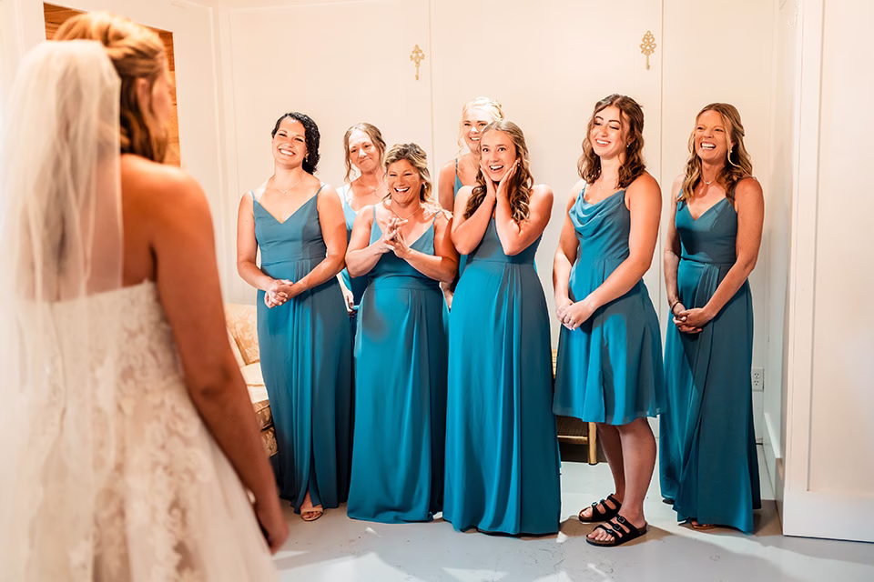 A bride in a wedding dress faces six bridesmaids in matching teal dresses who are smiling and reacting with joy in a well-lit room at a Barn at Copper Creek Wedding.