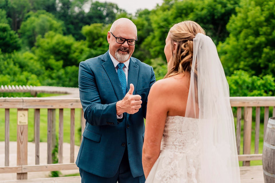 A man in a blue suit gives a thumbs up to a bride in a white dress and veil on a wooden deck with trees in the background at their Barn at Copper Creek Wedding.