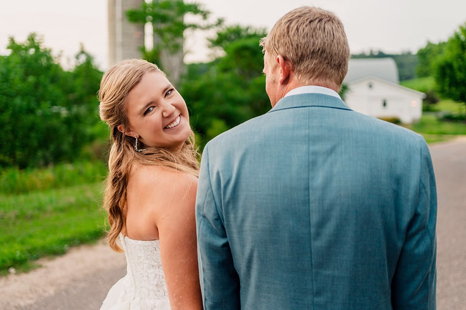 A bride in a white dress smiles at the camera while standing beside a man in a blue suit on a rural road, with greenery and a white house in the background at their Barn at Copper Creek Wedding.