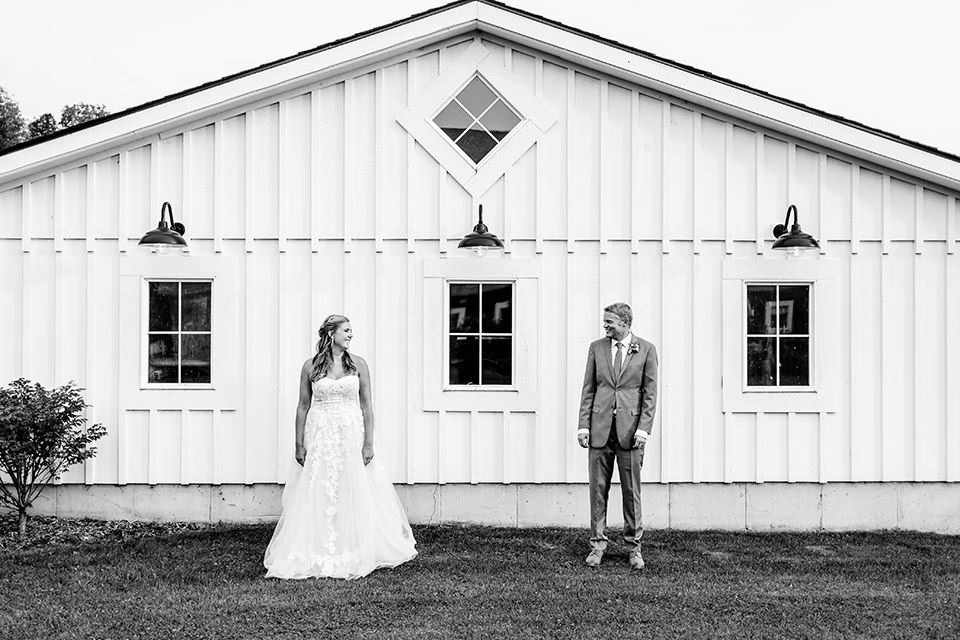 A bride and groom stand apart, facing each other in front of the white Barn at Copper Creek Wedding venue, featuring three windows and two light fixtures.