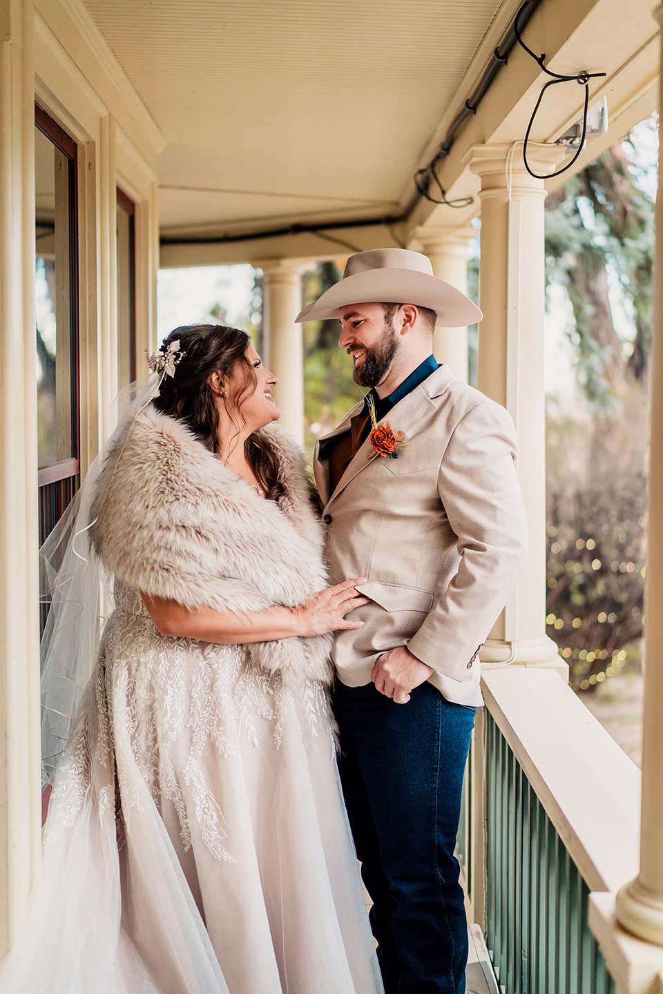 A bride in a lace gown and fur shawl stands beside a groom in a tan jacket and cowboy hat, smiling at each other on a porch.