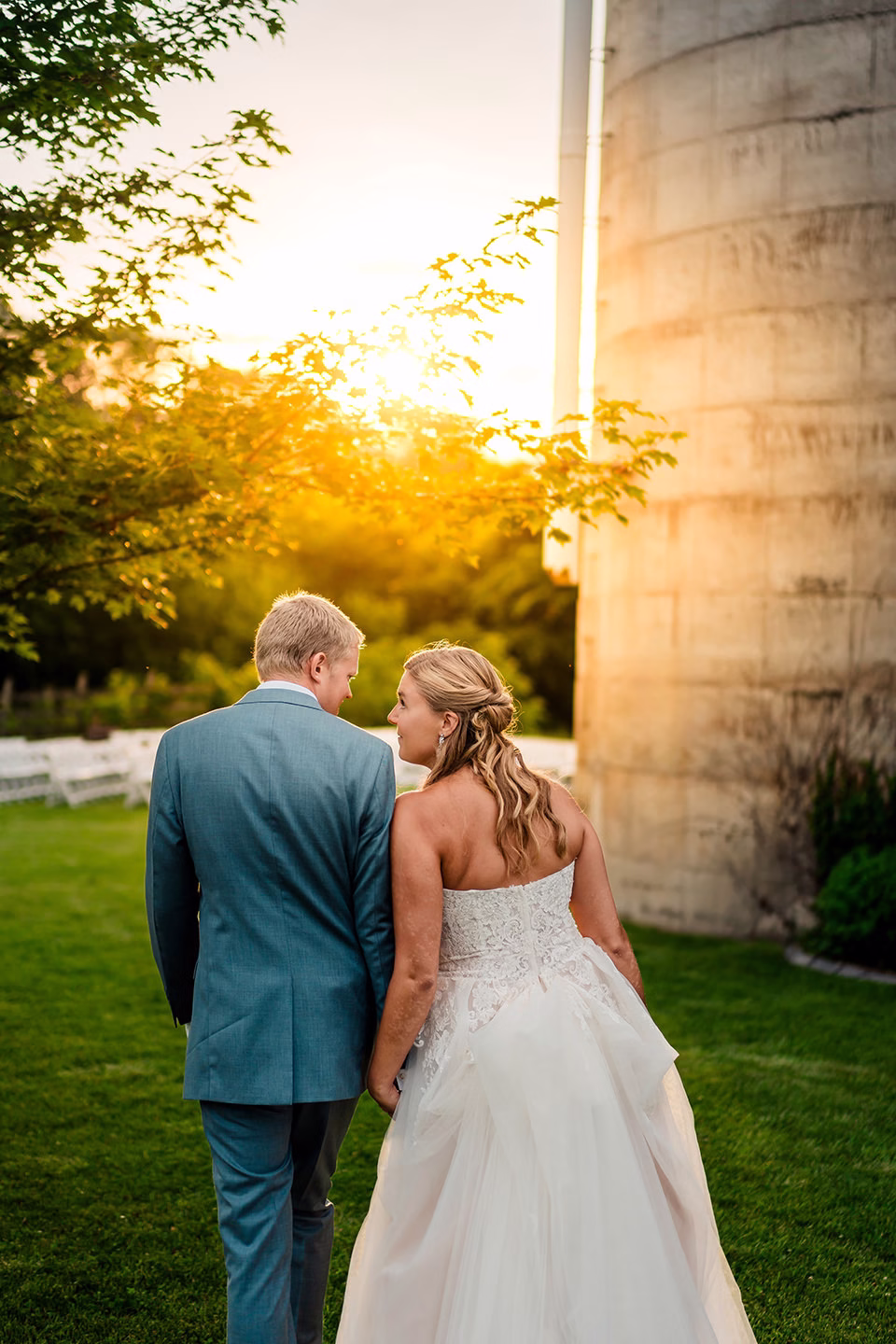 A bride and groom walk hand in hand outside at sunset, the bride in a white dress and the groom in a light blue suit, near a stone structure and green lawn during their Barn at Copper Creek Wedding.