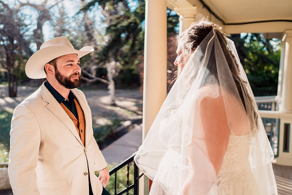 A man in a beige suit and cowboy hat stands facing a woman in a white wedding dress and veil on a porch in daylight.