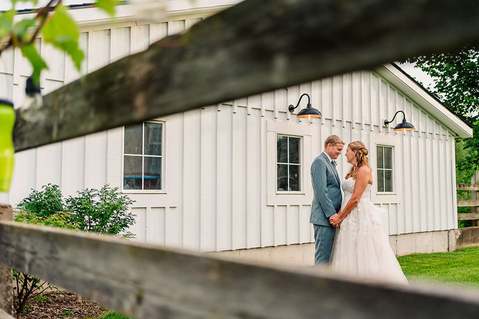 A bride and groom stand facing each other and holding hands outside a white barn at their Barn at Copper Creek Wedding, seen through the wooden slats of a fence.