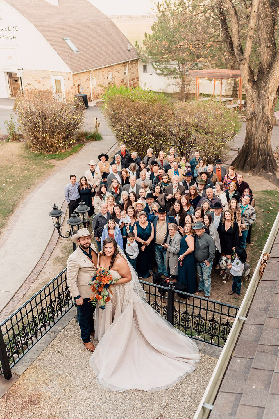 A bride and groom stand at the front while a large group of wedding guests pose together outside near a tree and a barn.