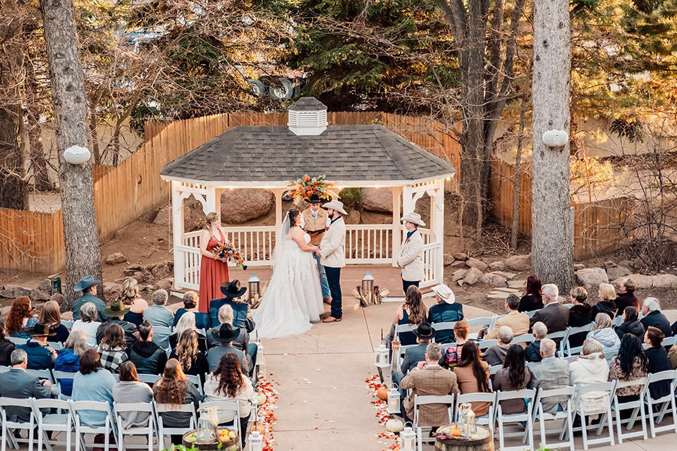 An outdoor wedding ceremony with a couple standing under a gazebo, surrounded by guests seated in rows on either side of a flower-petal-lined aisle.